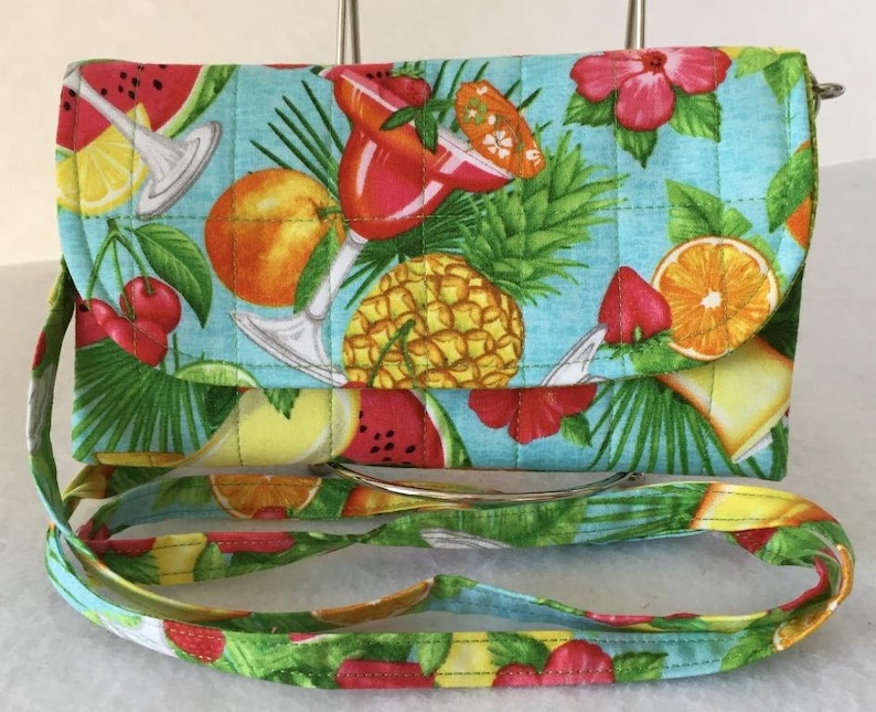 Pode incluir: Uma bolsa de ombro acolchoada turquesa com estampa de frutas tropicais. A estampa inclui abacaxi, melancia, laranjas, cerejas e uma ta&ccedil;a de margarita. A bolsa tem uma al&ccedil;a verde e amarela.
