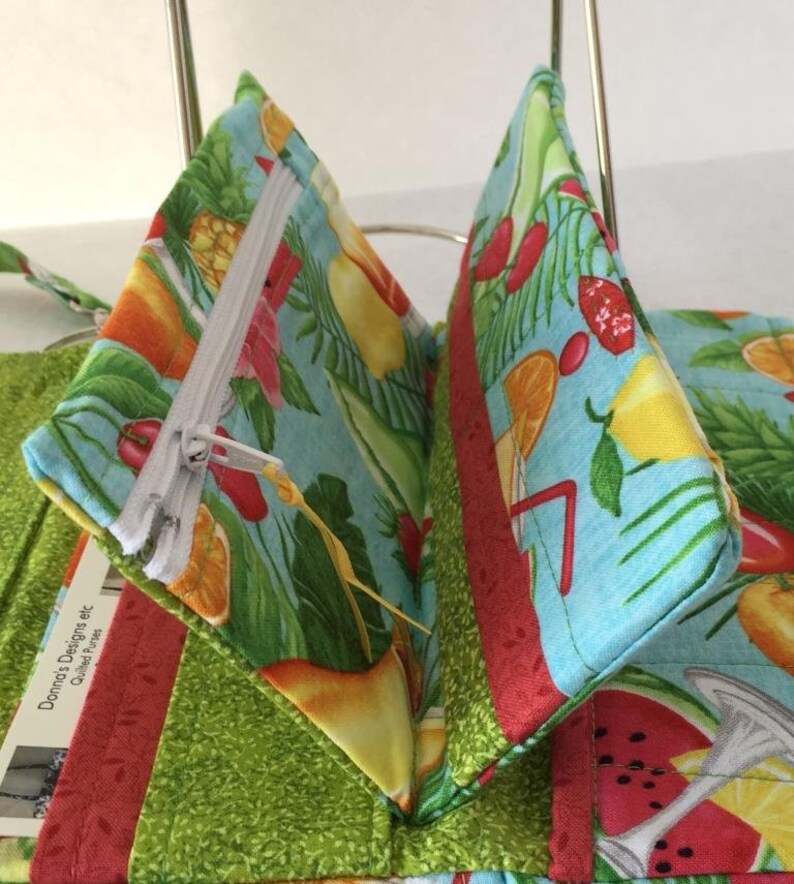 Pode incluir: Uma carteira de tecido turquesa com um bolso com z&iacute;per e uma estampa de frutas tropicais. A carteira est&aacute; aberta para mostrar o interior. O tecido &eacute; de algod&atilde;o e tem uma textura macia e acolchoada.