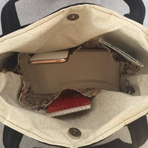 May include: A black tote bag with a beige fabric lining. The bag is open and shows a red notebook, a gold phone, and a white envelope inside.