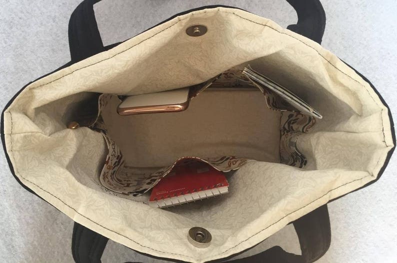 May include: A black tote bag with a beige fabric lining. The bag is open and shows a red notebook, a gold phone, and a white envelope inside.