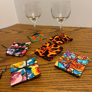 May include: Set of six colorful fabric coasters, each with a different pattern. The coasters are tied together with black ribbon and are stacked on a wooden table.