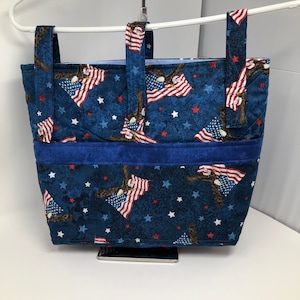 May include: A blue tote bag with a patriotic design featuring red, white, and blue American flags and bald eagles. The bag has a blue fabric lining and two blue straps.