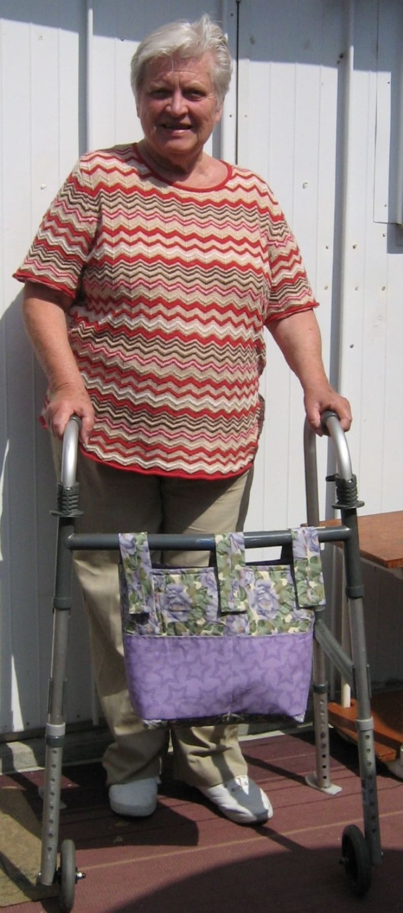 May include: A woman with white hair is using a walker with a floral patterned bag attached. She is wearing a red, white, and brown striped shirt and beige pants.