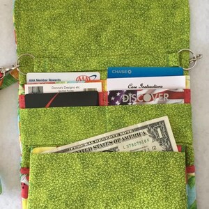 Pode incluir: Uma carteira de tecido verde com um design de estampa de frutas. A carteira tem v&aacute;rios bolsos para cart&otilde;es e dinheiro. A carteira tem um fecho de correr e uma correia de pulso.