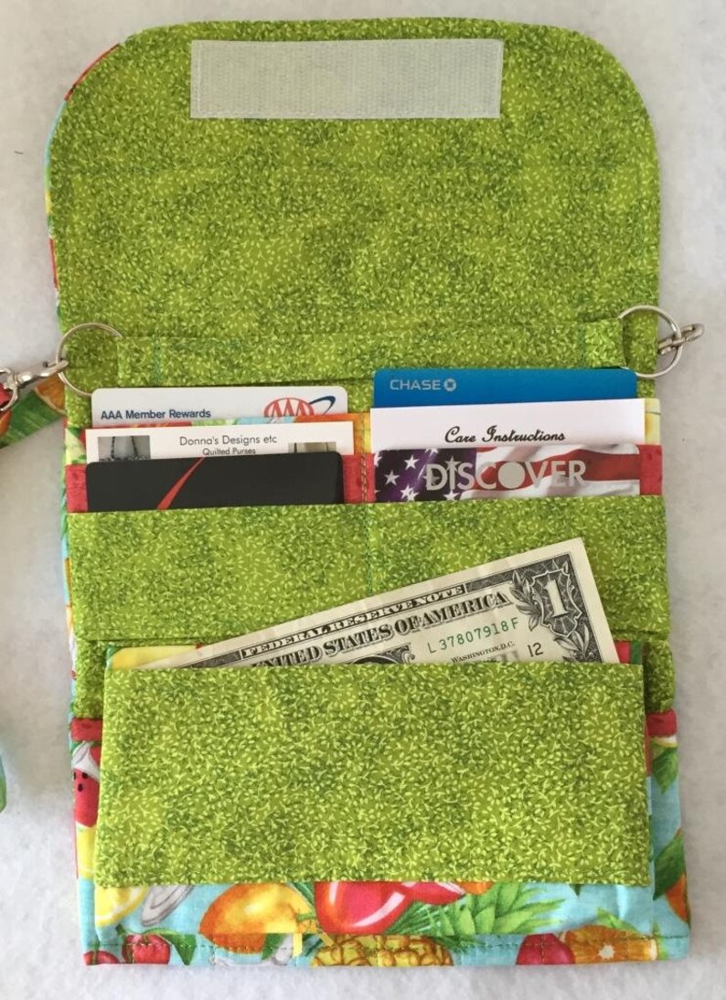 Pode incluir: Uma carteira de tecido verde com um design de estampa de frutas. A carteira tem v&aacute;rios bolsos para cart&otilde;es e dinheiro. A carteira tem um fecho de correr e uma correia de pulso.