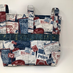 May include: A white fabric tote bag with blue and red accents. The bag features a repeating pattern of vintage Parisian postcards and stamps. The text "Paris" appears multiple times on the bag.