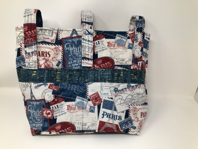 May include: A white fabric tote bag with blue and red accents. The bag features a repeating pattern of vintage Parisian postcards and stamps. The text "Paris" appears multiple times on the bag.
