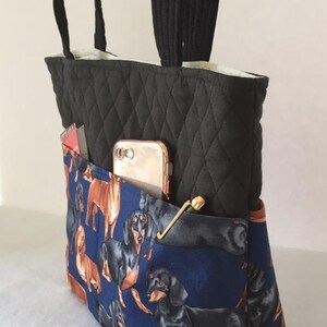 May include: Black quilted tote bag with a blue dachshund print pocket. The bag has black handles and a gold-colored clasp. A gold phone and a small notebook are visible in the pocket.