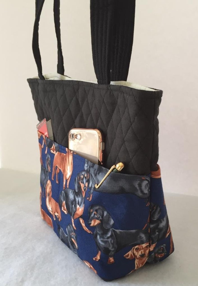 May include: Black quilted tote bag with a blue dachshund print pocket. The bag has black handles and a gold-colored clasp. A gold phone and a small notebook are visible in the pocket.
