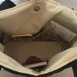 May include: The inside of a black tote bag with a beige fabric lining. The bag has multiple pockets and is empty except for a small notebook, a phone, and a piece of paper.