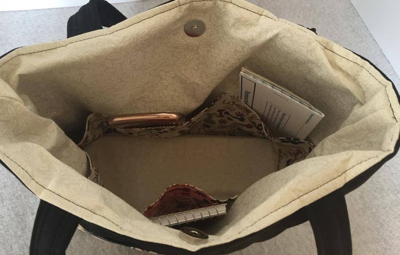 May include: The inside of a black tote bag with a beige fabric lining. The bag has multiple pockets and is empty except for a small notebook, a phone, and a piece of paper.