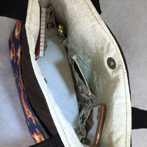 May include: Open view of a tote bag with a black exterior and handles. The interior features a floral pattern lining and several pockets. A notebook, pen, and phone are visible inside the bag. The bag has a magnetic snap closure.