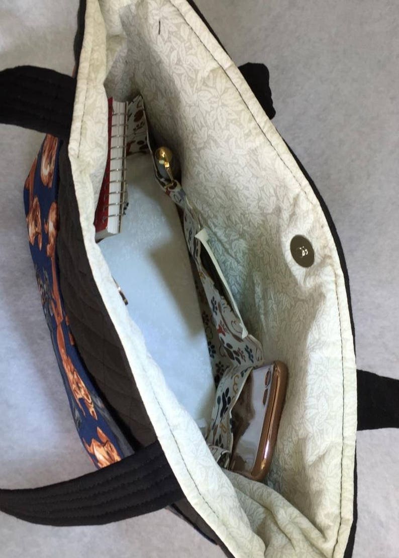 May include: Open view of a tote bag with a black exterior and handles. The interior features a floral pattern lining and several pockets. A notebook, pen, and phone are visible inside the bag. The bag has a magnetic snap closure.