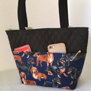 May include: Black quilted tote bag with a pocket featuring a dachshund print on a navy blue background. The bag has black handles and a light-colored interior. A smartphone, notepad, and a gold-colored pen are visible in the pockets.