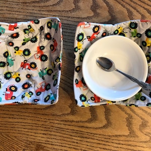 May include: Two white fabric bowl cozies with a tractor print. The cozies are folded and one has a white bowl and a silver spoon inside.