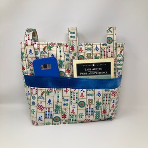 May include: A white tote bag with a mahjong tile pattern. The bag has two blue pockets and a blue strap. A blue phone and a book titled "Pride and Prejudice" by Jane Austen are inside the bag.