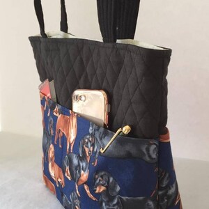 May include: Black quilted tote bag with black handles and a blue dachshund print pocket. The pocket holds a gold phone, a gold pin, and a red and white item. The bag is a stylish accessory.