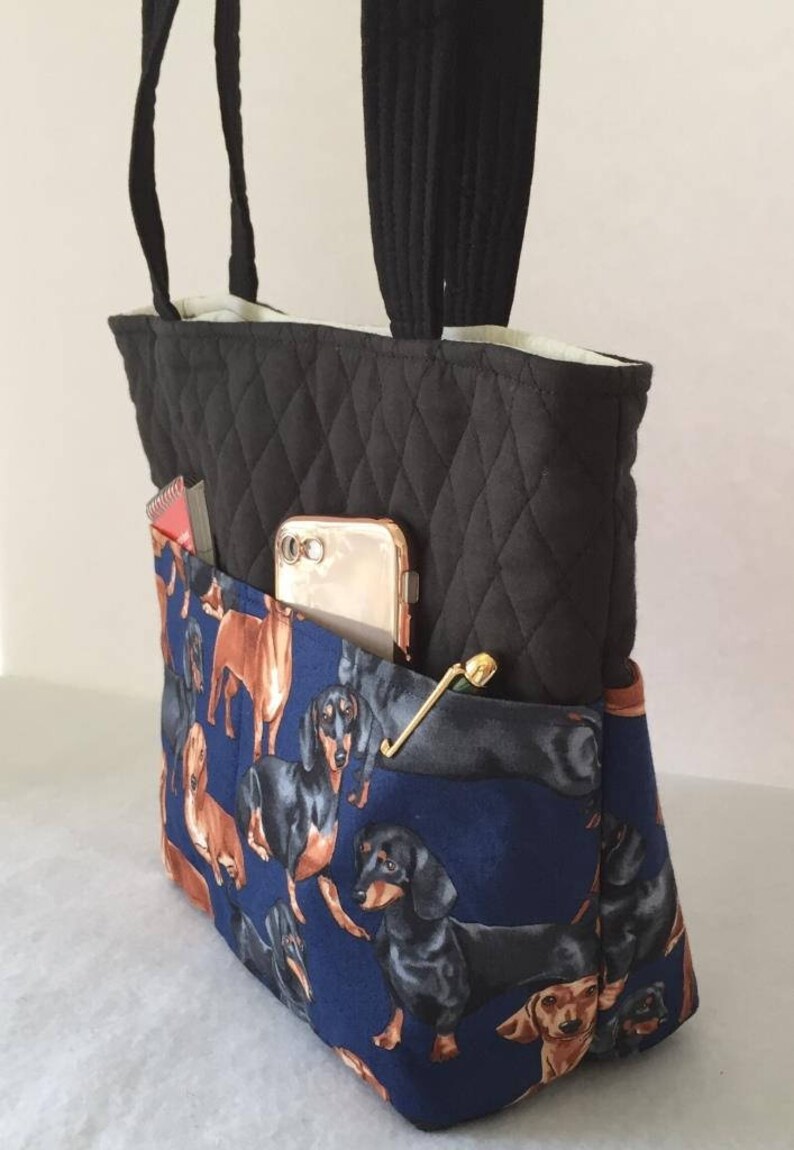 May include: Black quilted tote bag with black handles and a blue dachshund print pocket. The pocket holds a gold phone, a gold pin, and a red and white item. The bag is a stylish accessory.