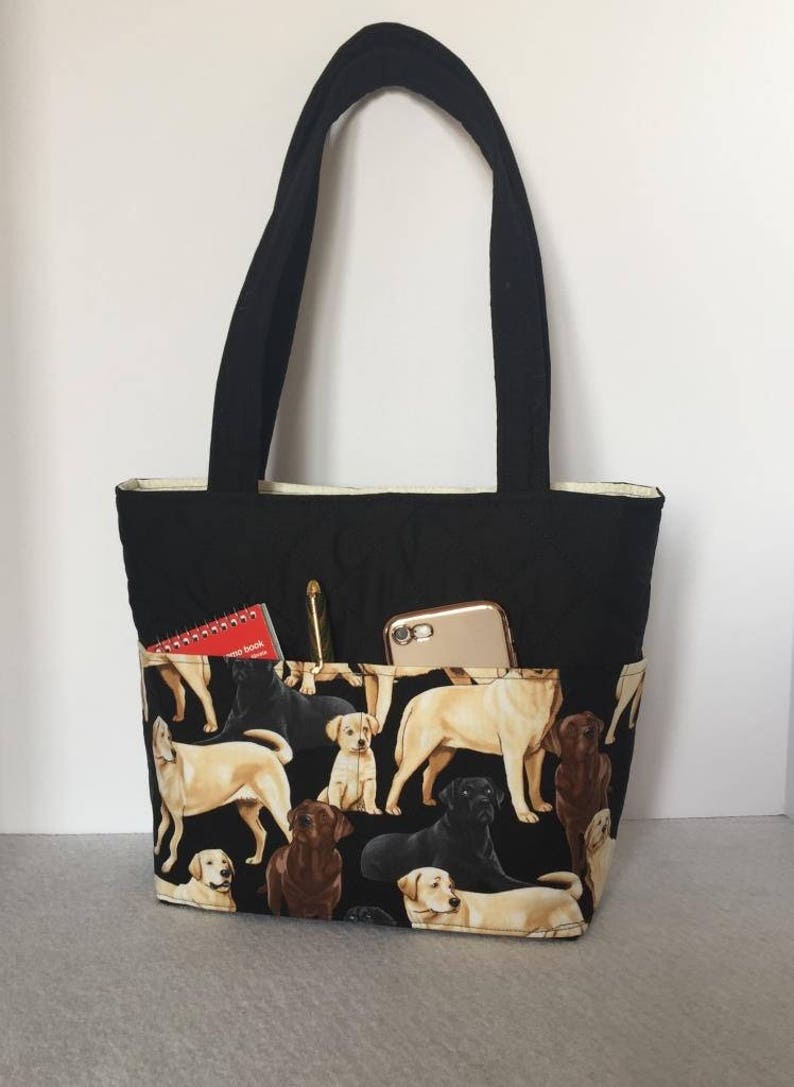 May include: Black tote bag with a zippered pocket and a phone pocket. The bag is lined with a cream fabric and has a pattern of Labrador Retrievers in black, brown, and yellow on the front.