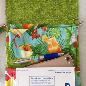 Pode incluir: Uma bolsa de tecido verde com estampa tropical e fecho de correr. A bolsa est&aacute; aberta e mostra um telem&oacute;vel, duas canetas e um registo de transa&ccedil;&otilde;es com o texto "Record your transactions" e "Transaction Register".