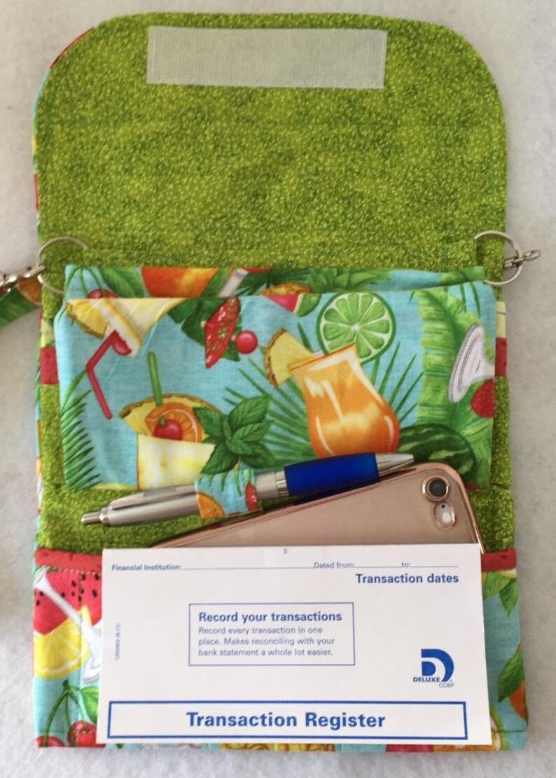 Pode incluir: Uma bolsa de tecido verde com estampa tropical e fecho de correr. A bolsa est&aacute; aberta e mostra um telem&oacute;vel, duas canetas e um registo de transa&ccedil;&otilde;es com o texto "Record your transactions" e "Transaction Register".