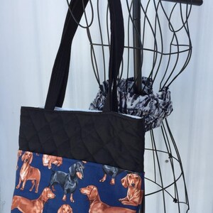 May include: Black quilted tote bag with long black straps. The bottom section features a blue fabric printed with dachshunds in various poses. A black and white patterned scarf is draped over a wire mannequin.