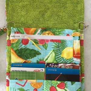Pode incluir: Uma carteira de tecido verde com estampa de frutas tropicais. A carteira tem um bolso com z&iacute;per, v&aacute;rias ranhuras para cart&otilde;es e um fecho de press&atilde;o. A carteira est&aacute; aberta e mostra uma variedade de cart&otilde;es de cr&eacute;dito no seu interior.