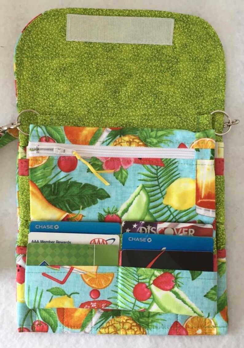 Pode incluir: Uma carteira de tecido verde com estampa de frutas tropicais. A carteira tem um bolso com z&iacute;per, v&aacute;rias ranhuras para cart&otilde;es e um fecho de press&atilde;o. A carteira est&aacute; aberta e mostra uma variedade de cart&otilde;es de cr&eacute;dito no seu interior.