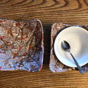May include: Two brown fabric bowl cozies with a mushroom print. The cozies are folded and ready to use. One cozy has a white bowl and a silver spoon inside.