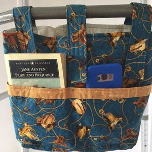 K&ouml;nnte beinhalten: Eine blaue Stofftasche mit einem Western-Motiv aus braunen S&auml;tteln und Seilen. Die Tasche hat zwei Taschen auf der Vorderseite, eine f&uuml;r ein Buch und eine f&uuml;r ein Telefon. Das Buch ist eine Penguin Classics Ausgabe von "Pride and Prejudice" von Jane Austen. Die Tasche h&auml;ngt an einem Metall-Rollator.