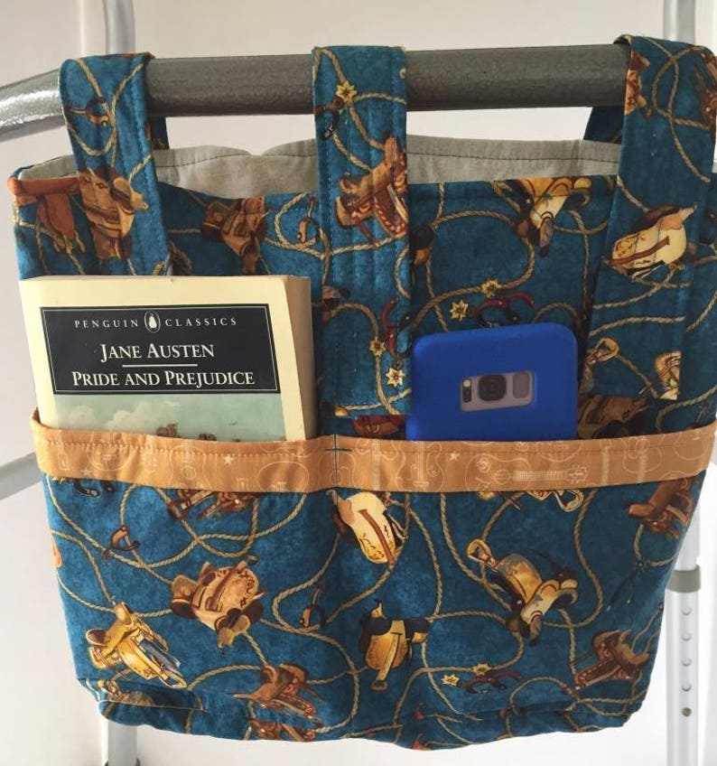 K&ouml;nnte beinhalten: Eine blaue Stofftasche mit einem Western-Motiv aus braunen S&auml;tteln und Seilen. Die Tasche hat zwei Taschen auf der Vorderseite, eine f&uuml;r ein Buch und eine f&uuml;r ein Telefon. Das Buch ist eine Penguin Classics Ausgabe von "Pride and Prejudice" von Jane Austen. Die Tasche h&auml;ngt an einem Metall-Rollator.
