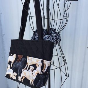 May include: Black quilted tote bag with a pocket featuring a print of golden retriever puppies. The bag has black straps and is hanging on a black wire rack.
