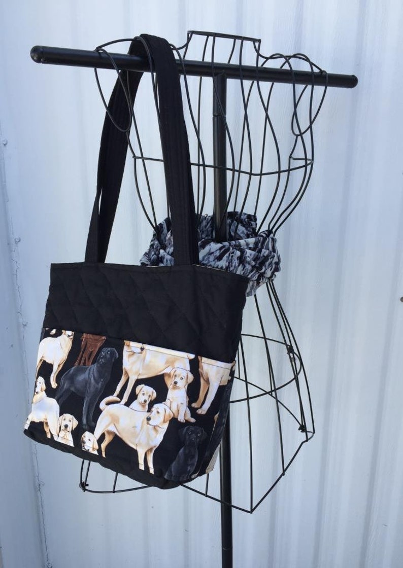 May include: Black quilted tote bag with a pocket featuring a print of golden retriever puppies. The bag has black straps and is hanging on a black wire rack.