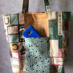 May include: A brown and beige patchwork tote bag with a blue phone case inside. The bag has a variety of patterns including horseshoes, guitars, and stars. The text "FAMILY LOVE COUNTRY LIFE" is visible on the bag.