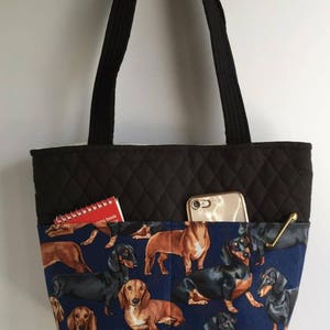 May include: A black quilted tote bag with black handles and a blue dachshund print pocket. The pocket contains a red notebook, a gold-colored pen, and a smartphone.