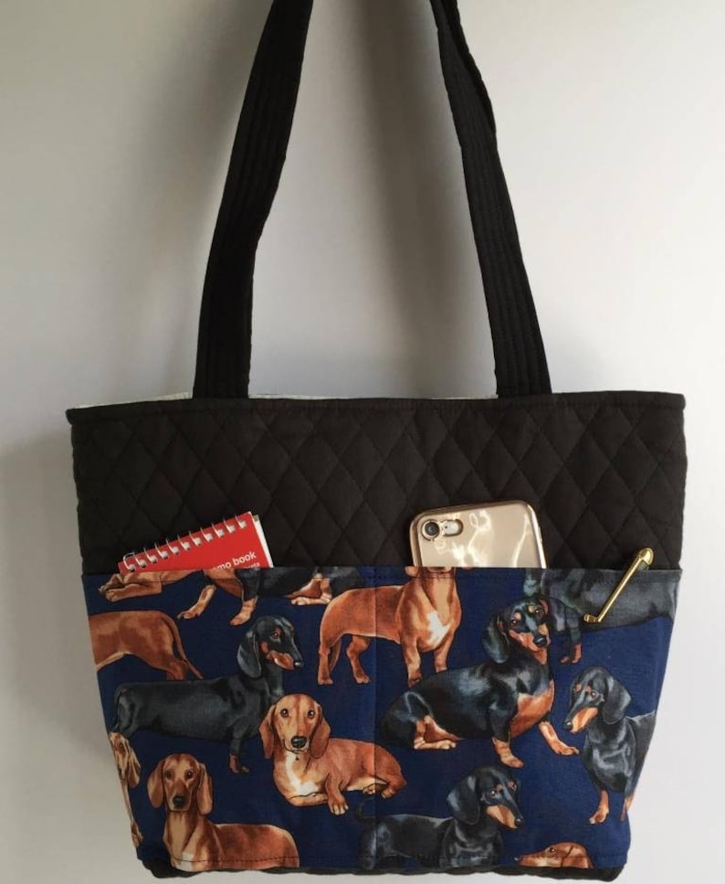 May include: A black quilted tote bag with black handles and a blue dachshund print pocket. The pocket contains a red notebook, a gold-colored pen, and a smartphone.
