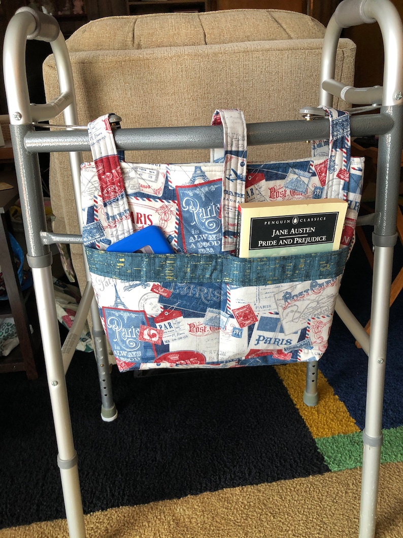 May include: A walker with a fabric bag hanging from the handles. The bag is decorated with a Parisian theme and has a book titled "Pride and Prejudice" by Jane Austen inside.