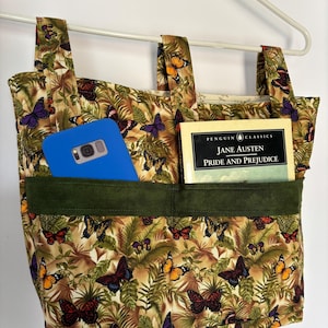 May include: A fabric hanging organizer with three straps, featuring a pattern of colorful butterflies and green foliage. It has a dark green horizontal pocket containing a blue smartphone and a copy of the book Pride and Prejudice by Jane Austen.