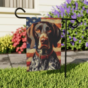 May include: A garden flag featuring a portrait of a German Shorthaired Pointer with a red collar, set against a backdrop of the American flag. The dog has brown and white speckled fur and is mounted on a black metal stand.