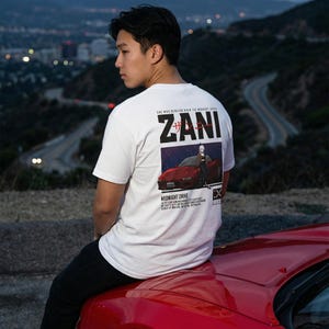 May include: White t-shirt with a graphic on the back featuring the word "ZANI" in black and red, a red car, and a character. The shirt also has the words "MIDNIGHT DRIVE" and other text. The person is sitting on a red car.