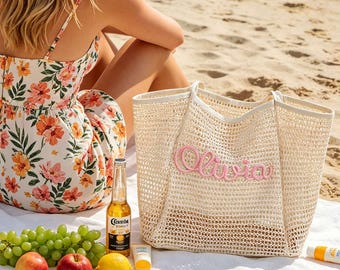 May include: A beach scene with a white woven tote bag embroidered with the name "Olivia" in pink. A floral dress, fruit, a beer bottle, and sunscreen are on a white towel. The scene is set on a sandy beach.