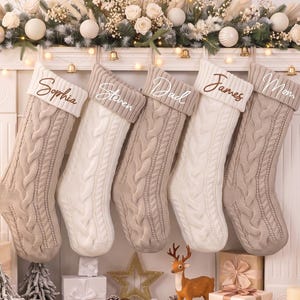 May include: Five cable-knit Christmas stockings hanging in a row, alternating between beige and white. Each stocking features a folded cuff embroidered with names: Sophia, Steven, Dad, James, and Mom in cursive script. The stockings are displayed against a white fireplace mantel decorated with greenery, white ornaments, and string lights.