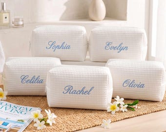 Puede incluir: Cinco neceseres rectangulares blancos con textura de gofre dispuestos sobre una alfombrilla tejida. Cada bolsa presenta un nombre bordado en letra cursiva azul claro: Sophia, Evelyn, Celilia, Rachel y Olivia. Cierre con cremallera.