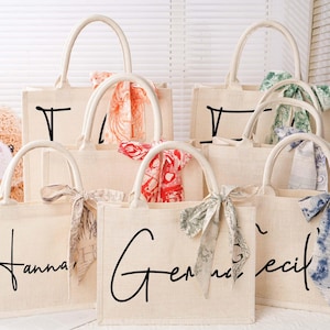 Peut inclure: Six sacs cabas de couleur naturelle avec des anses en tissu. Chaque sac porte un nom différent écrit en écriture noire sur le devant. Les sacs sont décorés de nœuds en tissu colorés.