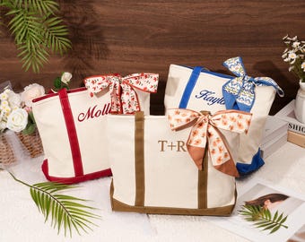 May include: Three personalized canvas tote bags with names and initials embroidered in red and brown. Each bag has a different colored trim and a decorative scarf tied to the handle. The bags are on a white surface.