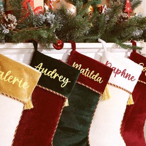 May include: Five personalised Christmas stockings in gold, dark green, red, and white velvet, each with a name embroidered in a complementary colour. Gold tassels and trim add detail. Festive holiday decor.