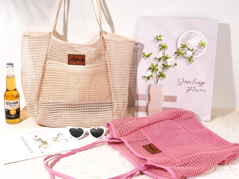 Pu&ograve; includere: Una borsa tote in rete beige e una borsa tote in rete rosa con etichette in pelle. Sono presenti anche una bottiglia di birra Corona, occhiali da sole a forma di cuore e una stampa floreale decorativa. La stampa floreale recita "Smiling Flowers".