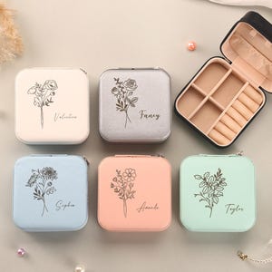 Puede incluir: Una colección de joyeros cuadrados en varios colores pastel, incluyendo blanco, plateado, azul, melocotón y verde menta. Cada estuche presenta un diseño floral y un nombre. Un estuche está abierto, revelando compartimentos para joyas.