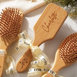 以下が含まれることがあります： 明るい茶色の毛と竹の柄が付いた木製のヘアブラシ。ブラシの1つには「Charlotte」という名前が刻まれています。ブラシは「Just For You」と書かれたクリーム色のリボンで結ばれています。