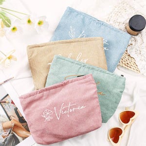Puede incluir: Cuatro neceseres de maquillaje de pana en colores pastel: azul claro, beige, verde menta y rosa. Cada neceser tiene texto bordado en blanco y un diseño floral. Los neceseres están colocados sobre una superficie blanca.
